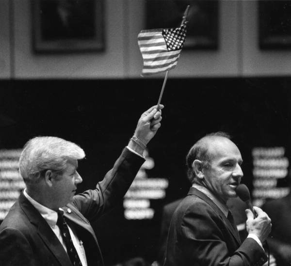 State Senator Dick Langley of Clermont argues against metric conversion, claiming that only drug dealers and communists would support the bill because drugs were sold in metric units and communist countries used the metric system. Senator George Kirkpatrick waves a small American flag over Langley's head (1984).