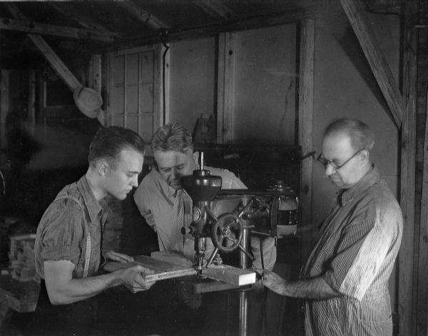 A student feeds a piece of wood through a router as his instructors look on (photo circa 1936).