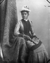 Seated woman wearing sunflower hat, holding a fan.